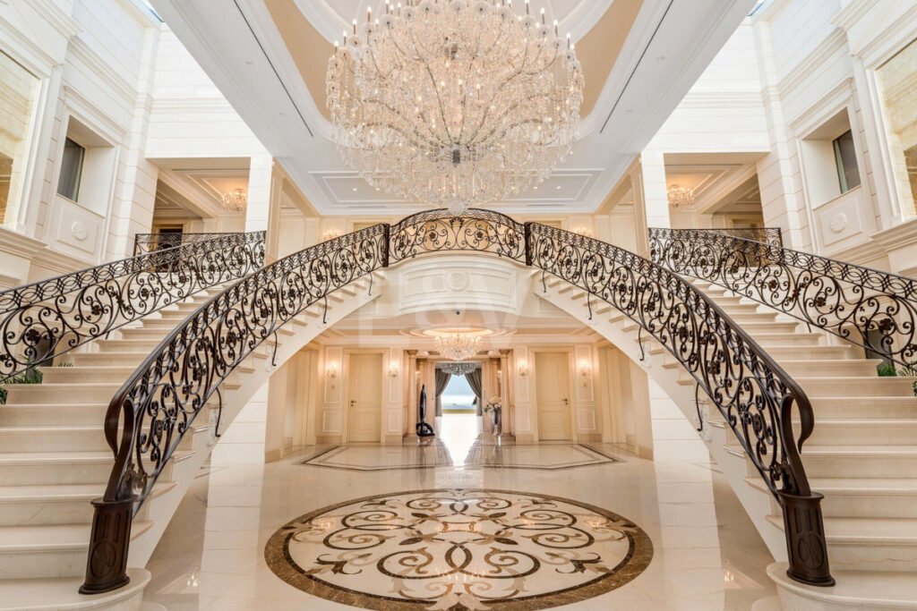 Impresionante escalera doble con barandillas forjadas y gran lámpara de araña en el vestíbulo principal de Villa Bellagio.