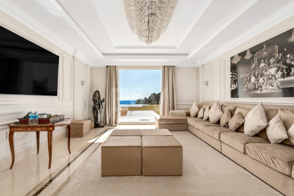 Salón en Villa Bellagio con sofá en L, arte mural, lámpara de cristal y vistas al mar desde la terraza.