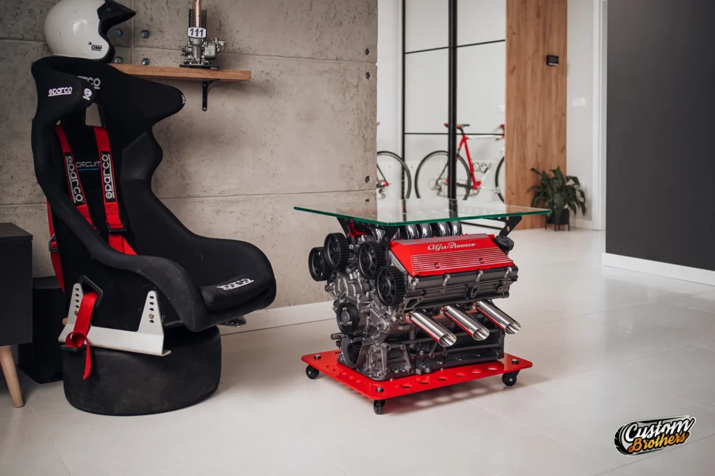 Mesa de diseño con motor Alfa Romeo V6 y cristal, junto a pared con trofeos y decoración automovilística