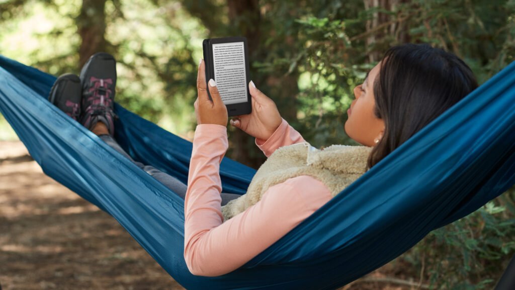 Mujer relajándose en una hamaca azul mientras lee un Kindle Paperwhite en un entorno natural con árboles.