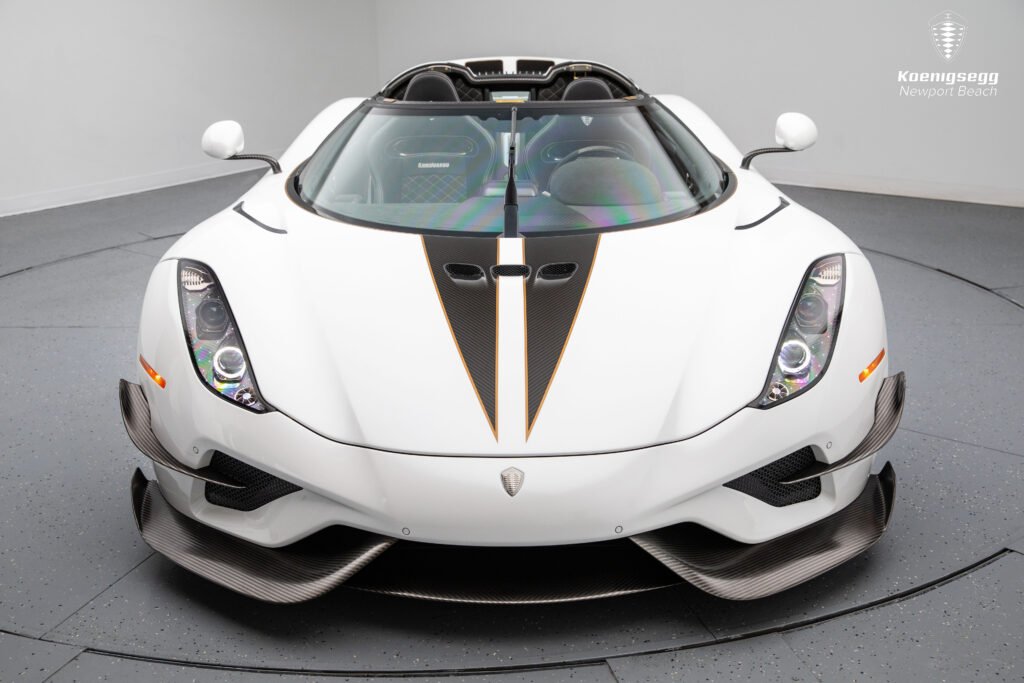 Vista frontal del Koenigsegg Regera 2019 en blanco con detalles en fibra de carbono y líneas doradas, resaltando su diseño aerodinámico